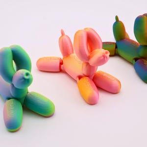 May include: Three colorful balloon dog sculptures in shades of pink, orange, green, and blue. The whimsical figures are posed in different positions, with one lying down and two standing. The dogs are on a white background.