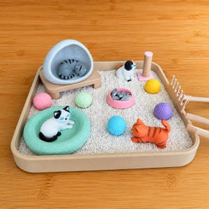 May include: A square beige tray filled with white sand containing a miniature cat-themed zen garden. It features a gray cat in a stone-like cave, a white cat in a mint green bed, an orange tabby cat, a scratching post, a pink food bowl, and colorful yarn balls.