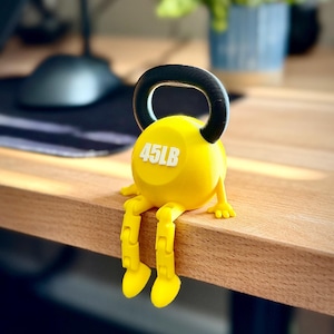 May include: A yellow novelty kettlebell with the text "45LB" printed on the front. The kettlebell has a black handle and yellow legs and feet, giving it a cartoon-like appearance. It is sitting on a wooden surface.