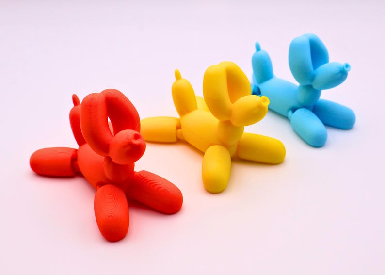 May include: Three colorful balloon dog sculptures in red, yellow, and blue. The whimsical figures are arranged on a white surface. Each dog has a distinct shape, resembling the classic balloon animal design.