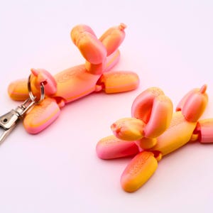 May include: Two orange and pink balloon dog keychains with silver hardware. The keychains are in the shape of a dog, with articulated joints. One keychain has a silver clasp attached.