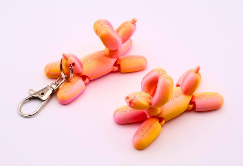 May include: Two orange and pink balloon dog keychains with silver hardware. The keychains are in the shape of a dog, with articulated joints. One keychain has a silver clasp attached.