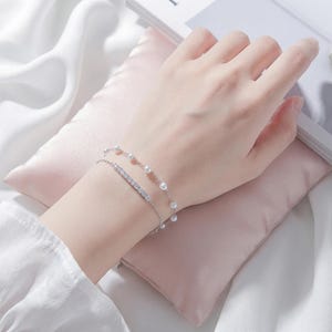 以下が含まれることがあります： 銀色の二連ブレスレット。細いチェーンに小さな白い球状のパールが等間隔に並んだものと、透明で輝く長方形のカットストーンが一列に並んだチェーンの二本で構成されています。