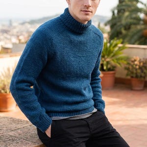 May include: A long-sleeved, deep blue knit sweater featuring a ribbed turtleneck collar, ribbed cuffs, and a ribbed hem. The sweater is worn over a simple grey undershirt, paired with solid black trousers, showcasing a textured, wool-like knit pattern.