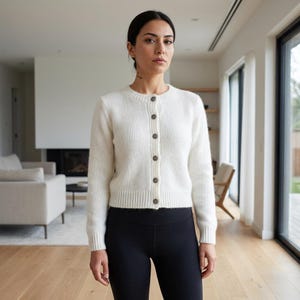 May include: A white long-sleeved knit cardigan featuring a crew neckline and a front button-down closure with six dark circular buttons. The garment has a ribbed hem and ribbed cuffs, constructed from a soft, textured material.