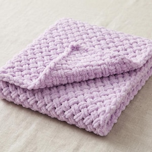 Könnte beinhalten: Eine gefaltete, hellviolette Decke mit einer groben, gewebten Korbgeflechtstruktur. Das Material wirkt weich und flauschig, wobei dicke, miteinander verbundene Schlaufen ein gleichmäßiges, sich wiederholendes Muster auf der gesamten Oberfläche bilden.