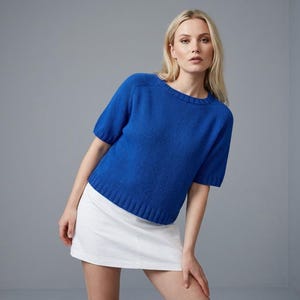May include: A vibrant royal blue short-sleeved knit sweater featuring a ribbed crew neckline, ribbed sleeve cuffs, and a ribbed hemline, paired with a plain white mini skirt. The sweater has a classic pullover construction with a textured knit finish.