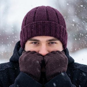 Op de afbeelding: Een donkerpaarse, geribbelde gebreide muts gecombineerd met donkerbruine gebreide handschoenen en een zwarte jas met capuchon. De accessoires worden gedragen tijdens een sneeuwbui, met zichtbare sneeuwvlokken op de donkere stof van de kleding.