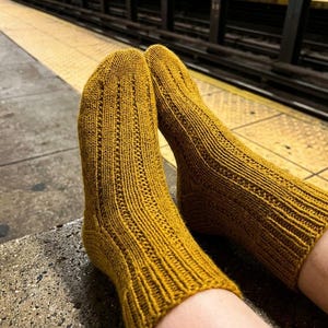May include: A pair of mustard yellow knit socks featuring a vertical ribbed texture with small eyelet details running along the length of the foot and ankle. The socks are crafted from a textured yarn and have a fitted heel and cuff construction.