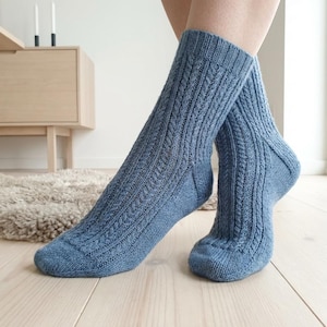Puede incluir: Un par de calcetines de punto a media pantorrilla en color azul polvoriento. Los calcetines presentan un patrón de punto de ochos vertical a lo largo de la pierna y el pie, con una textura de punto liso en las secciones de la puntera y el talón.