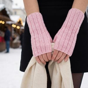Op de afbeelding: Een paar lichtroze vingerloze handschoenen met een verticaal geribbeld breipatroon. De handschoenen worden over de handen gedragen en reiken tot voorbij de polsen, gecombineerd met een effen crèmekleurige sjaal voor een zwarte rok.