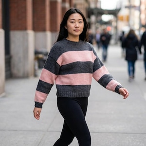 May include: A long-sleeve knit jumper featuring wide horizontal stripes in alternating dark charcoal grey and soft pastel pink. The garment has a classic crew neckline, ribbed cuffs, and a ribbed hem, constructed with a chunky yarn texture.