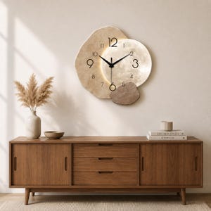 Può includere: Un orologio da parete con una cornice beige irregolare e testurizzata e un centro circolare illuminato che ricorda la luna. Il quadrante presenta numeri arabi neri e lancette sottili nere. Un pezzo marrone testurizzato è fissato alla struttura.