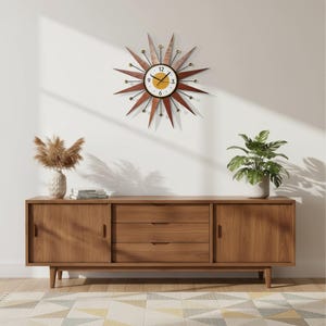 Pode incluir: Um relógio de parede em forma de estrela com mostrador branco, centro amarelo e números pretos, apresentando longos raios de madeira e finos raios de metal preto com pequenas esferas douradas nas pontas, montado sobre um aparador de madeira.