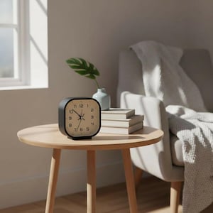 May include: A square-shaped analog alarm clock with a black frame and a white face featuring black numerals from 1 to 12. The clock has thin black hour, minute, and second hands, and it sits on a flat surface next to a stack of three books.