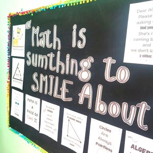May include: A black bulletin board with a colorful pom-pom border featuring the text "Math is sumthing to SMILE About" in large white letters. Several white paper signs with math-themed jokes and illustrations are pinned to the board surface.