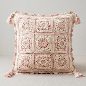 Op de afbeelding: Een vierkante gehaakte kussenhoes met een raster van negen granny squares in crème en zachtroze garen. Het ontwerp is voorzien van een geschulpte rand en vier bijpassende kwastjes op de hoeken, met een klein knoopje aan de zijkant.