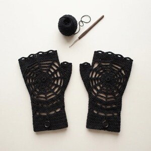 May include: A pair of black fingerless gloves featuring a crocheted spider web pattern, each with a single button at the wrist. A ball of black yarn and a metal crochet hook are positioned above the gloves on a plain, light-colored surface.