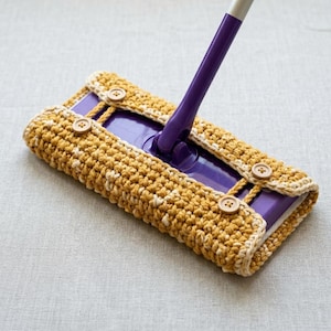 May include: A purple flat mop head fitted with a removable, textured mustard yellow and cream crocheted cover. The cover is secured around the mop base with four small, round, light brown buttons, featuring a visible handle attachment point.