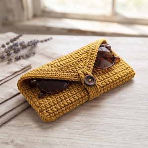 May include: A mustard yellow crocheted case with a triangular flap secured by a single dark brown button. The case contains a pair of dark-rimmed sunglasses with brown lenses, which are partially visible through the opening of the textured pouch.