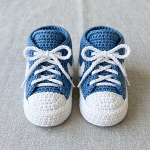 May include: A pair of handmade baby sneakers in a classic high-top style. The shoes are crocheted with blue yarn for the uppers and white yarn for the toe caps and laces. The white laces are tied in bows.
