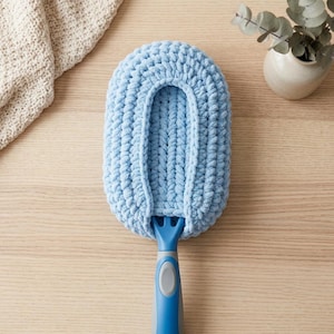 May include: A light blue oval-shaped duster head made of thick, chunky yarn, featuring a textured crochet-style knit pattern. The duster head is attached to a blue and gray plastic handle with a smooth, ergonomic grip and a central thumb indentation.