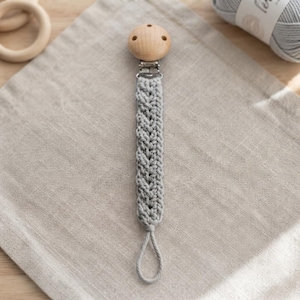 May include: A pacifier clip featuring a light gray crocheted strap with a braided texture. The strap is attached to a round, natural-colored wooden clip at the top and ends with a matching gray yarn loop at the bottom for securing a pacifier.