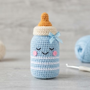 Puede incluir: Un juguete de biberón tejido a ganchillo con rayas azul claro y blancas. El biberón presenta una tetina color canela, una tapa color crema y un pequeño lazo de cinta azul claro. Tiene una cara bordada con ojos cerrados y mejillas rosas.