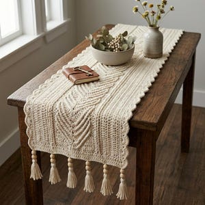 May include: Cream-colored crochet table runner with a geometric pattern and scalloped edges. The runner has tassels with wooden beads at the ends. A small leather-bound journal and a vase with flowers sit on the wooden table.