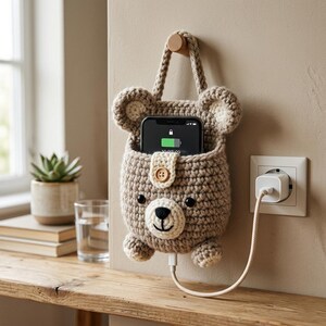 Könnte beinhalten: Eine beige gehäkelte Handy-Ladetasche in Bärenform mit runden Ohren, cremefarbener Schnauze, schwarzen Augen und einem kleinen Holzknopf. Die Tasche hängt an einem Wandhaken und enthält ein Smartphone mit grünem Akkusymbol.