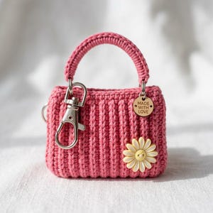 May include: A small, pink crocheted handbag keychain with a silver-tone clasp and handle. A wooden tag reads "MADE WITH LOVE." A yellow and white daisy is attached to the front of the bag. The bag is approximately 3 inches wide.