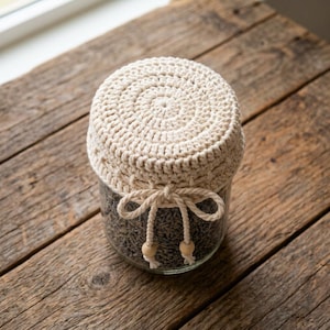 May include: A clear glass jar filled with dried lavender buds, topped with a cream-colored crocheted cover. The cover features a matching cream-colored cord tied in a bow around the neck, with two small wooden beads attached to the cord ends.