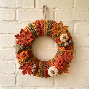 May include: A circular crochet wreath featuring alternating vertical segments of orange, yellow, brown, and green yarn. The wreath is decorated with crocheted maple leaves in orange and brown, small pumpkins in orange and cream, and brown acorns.