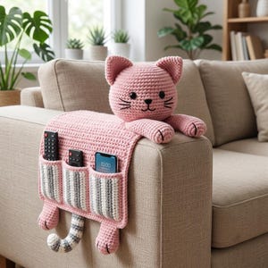 May include: A pink crocheted cat-shaped sofa armrest organizer featuring a head with black eyes and whiskers, two front paws, and a striped tail. The main body includes three vertical pockets with grey and white stripes holding two remotes and a smartphone.