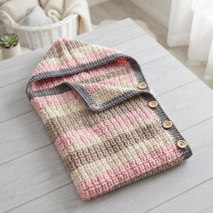 May include: A hooded crochet blanket featuring horizontal stripes in shades of pink, cream, and brown. The edges are trimmed in dark gray yarn, and the side is secured with four round, light brown wooden buttons along the folded flap border.