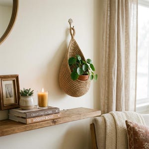 May include: A hanging plant holder, woven in a natural tan hue, suspended from a wall hook. It cradles a potted plant with green foliage. The scene includes a wooden shelf with books, a candle, and a framed picture, creating a relaxed, bohemian atmosphere.