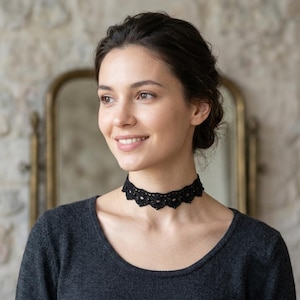 Può includere: Una collana girocollo in pizzo nero caratterizzata da un design con bordo smerlato, intricati motivi intrecciati e piccole perline incastonate, indossata sopra un top a maniche lunghe grigio scuro con scollo rotondo.
