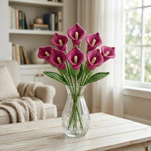 May include: A bouquet of crocheted calla lilies featuring vibrant magenta funnel-shaped petals and cream-colored spadices. The flowers are attached to long green stems with textured leaves and are arranged in a clear, faceted glass vase.