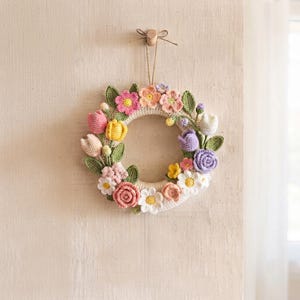 Puede incluir: Una corona circular de ganchillo con una variedad de flores coloridas, incluyendo tulipanes, rosas y margaritas en tonos rosa, amarillo, morado y blanco con hojas verdes. La corona está colgada de un cordel fino en un pequeño gancho de pared.
