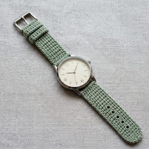 Op de afbeelding: Een polshorloge met een ronde zilverkleurige kast, een crèmekleurige wijzerplaat en dunne wijzers. Het horloge is bevestigd aan een saliegroene, gehaakte horlogeband met een zilverkleurige metalen gesp en meerdere gaatjes voor een goede pasvorm.