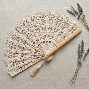 May include: A hand fan featuring a beige crochet lace canopy with an intricate floral pattern and a scalloped edge. The fan is attached to a light-coloured wooden handle and includes a matching beige tassel hanging from the base pivot point.
