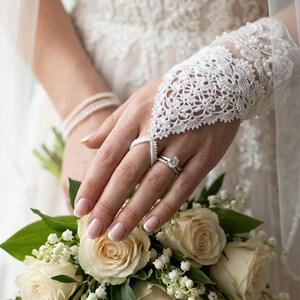 May include: A white lace hand flower bracelet featuring intricate floral patterns and small bead accents. The accessory connects to a finger loop and covers the back of the hand, worn with a silver ring featuring a round center stone.