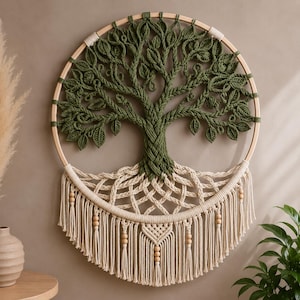 Op de afbeelding: Een ronde macramé wandhanger met een boomontwerp van olijfgroene koordbladeren en takken. De stam gaat over in een crèmekleurig wortelstelsel, geweven met ingewikkelde knopen en houten kralen, afgewerkt met lange franjes en kwastjes.
