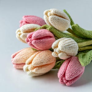 May include: A bouquet of crocheted tulips in shades of cream, soft pink, and peach. Each flower features a textured, knitted petal structure with green crocheted stems and leaves attached to the base of the blooms, creating a floral arrangement.