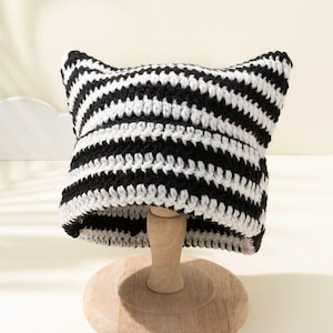 May include: A crochet beanie featuring a black and white horizontal striped pattern. The hat has two pointed corners at the top, creating a cat-ear silhouette. The knit texture is visible throughout the piece, showcasing the alternating yarn colors.