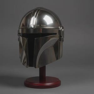 May include: A polished silver-coloured helmet with a smooth, reflective surface and a T-shaped visor opening. The helmet features a central ridge running from the crown to the brow and is mounted on a dark red circular pedestal stand.