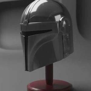 May include: A metallic silver helmet with a T-shaped visor opening, featuring a smooth, reflective surface with subtle scuff marks. The helmet is mounted on a dark red circular base with a vertical support post, displaying a sleek, curved design.