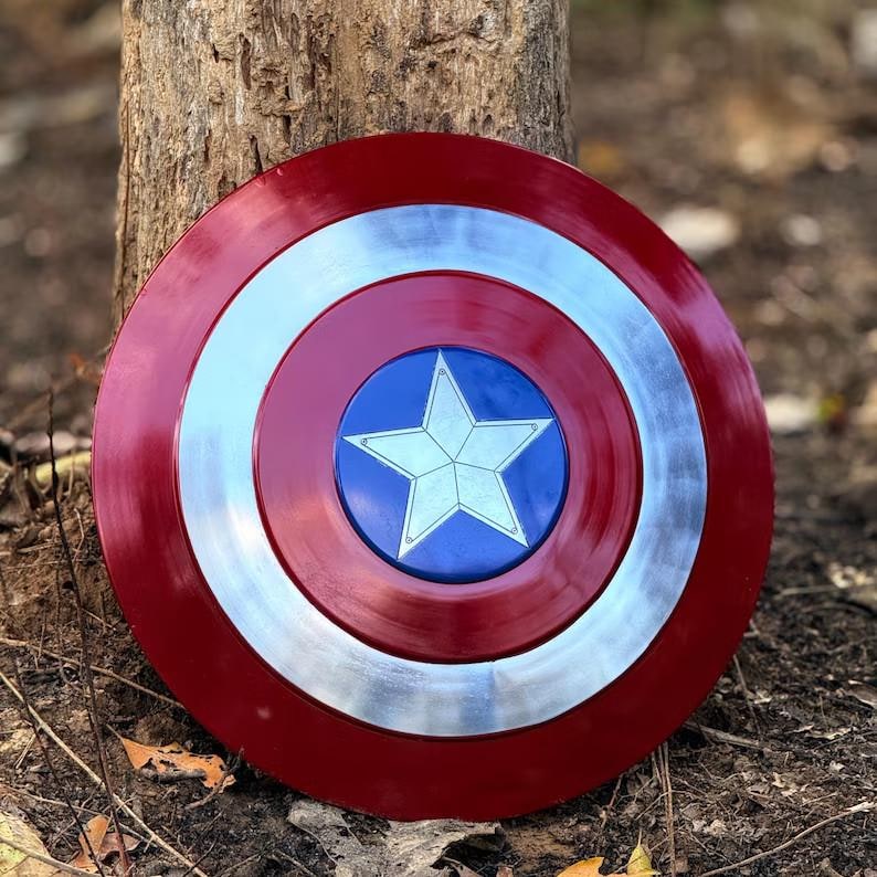 Op de afbeelding: Een rond schild in de kleuren van Captain America. Het heeft een rode buitenring, een zilveren ring en een blauw midden met een witte ster. Het schild is van metaal en ligt op een natuurlijke achtergrond.