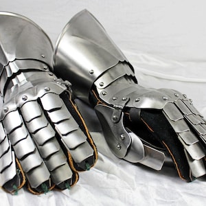 May include: A pair of articulated steel gauntlets featuring overlapping metal plates covering the fingers and back of the hand, attached to a solid cuff, with a black fabric lining visible at the interior and along the finger segments.
