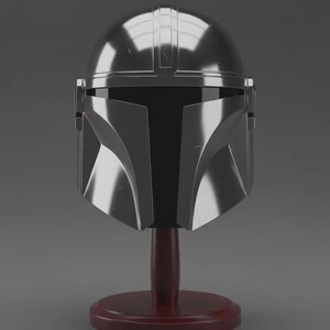 May include: A polished silver-coloured helmet with a smooth, rounded dome and a distinct T-shaped black visor section. The helmet features a central ridge and angular cheek plates, mounted on a dark reddish-brown circular wooden display stand.